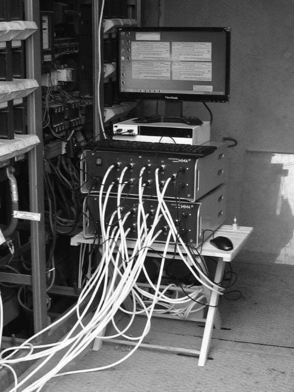 Two rack-mounted MM4 measuring units, a desktop computer and a monitor standing on a wooden trestle table inside a distribution substation, with eight bundles of white cable running out of them into the cabinet alongside.